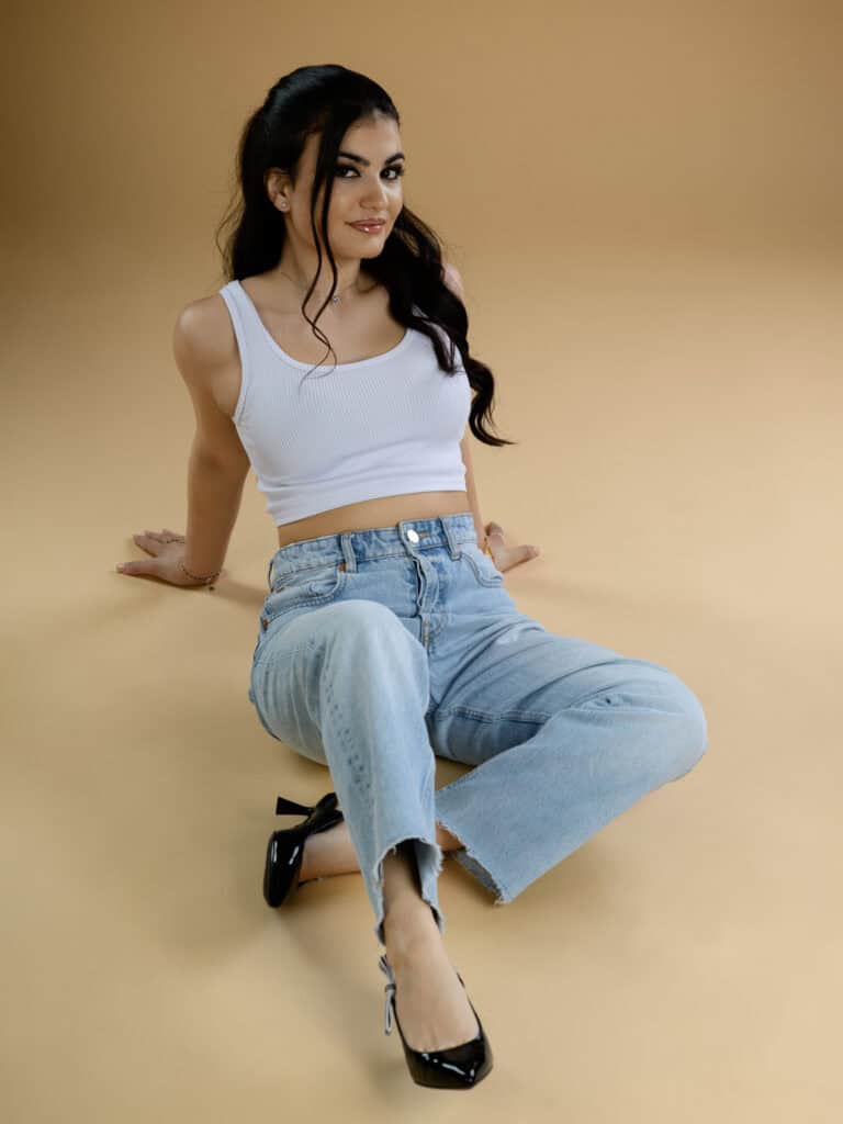 Jeune femme mode en jeans et top blanc, assise sur un fond beige, look décontracté.