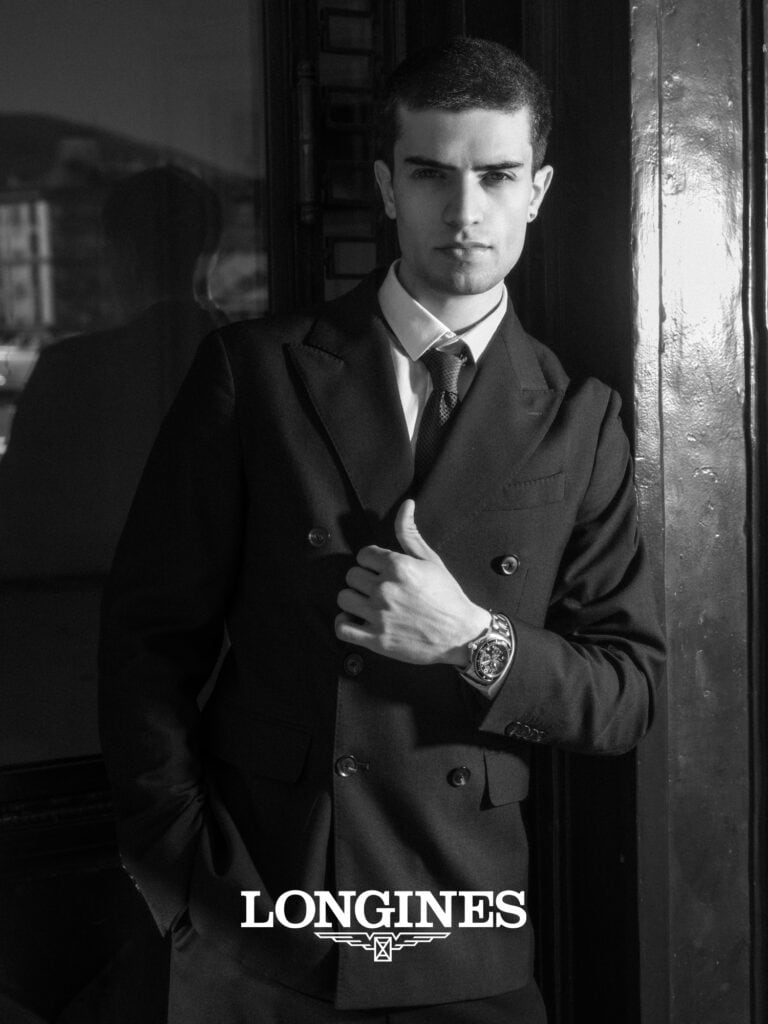 Homme en costume noir sophistiqué, regard sérieux, dans un environnement urbain.