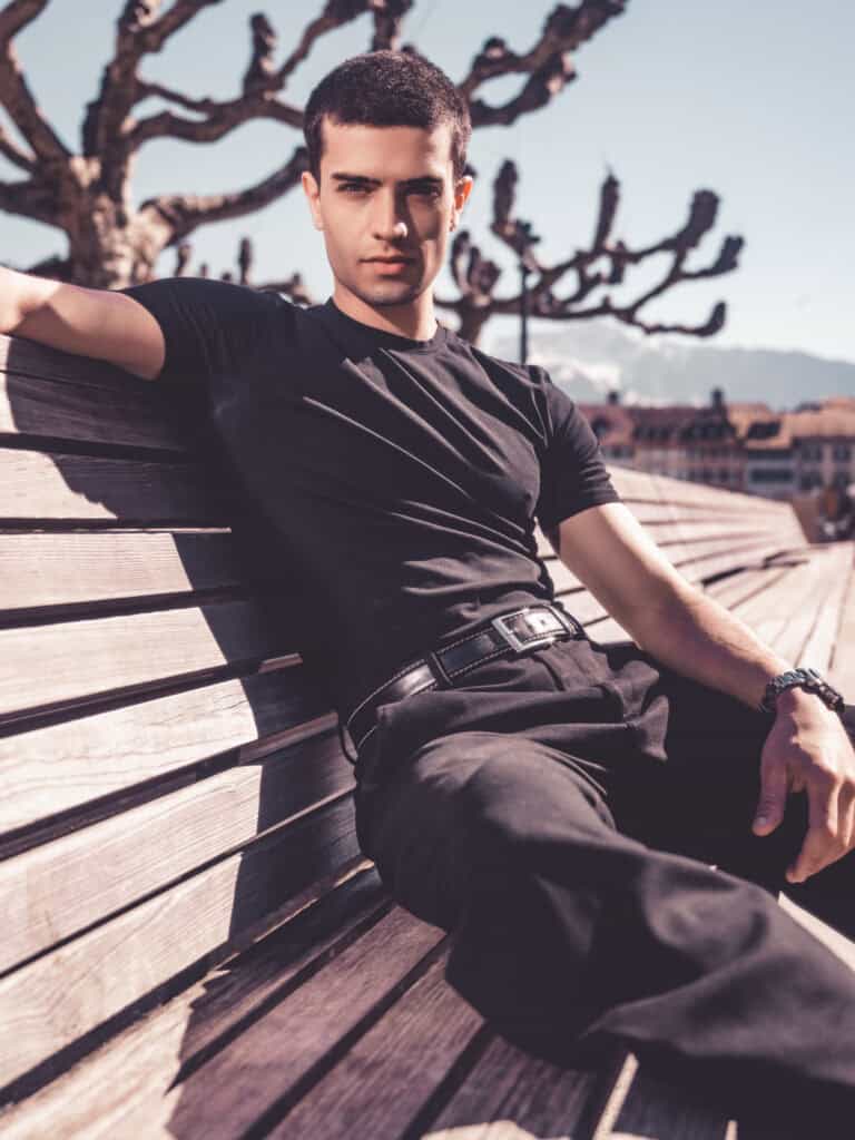 Homme jeune assis détendu sur un banc en bois dans un parc urbain, paysage urbain en arrière-plan, s.