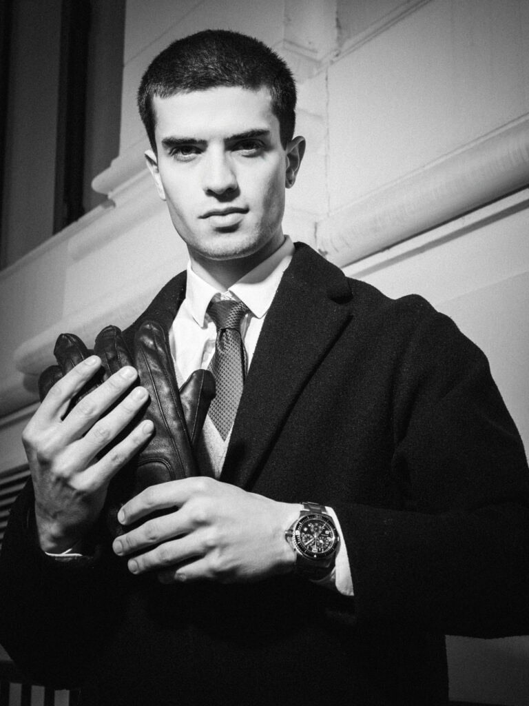 Vieux homme en costume avec gants en cuir, regard sérieux, dans un cadre urbain noir et blanc.