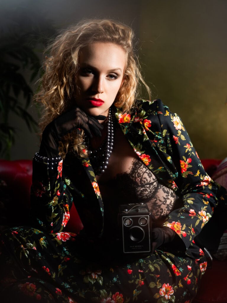 Femme sophistiquée en tenue florale et lingerie noire, posant avec un appareil photo dans un studio.