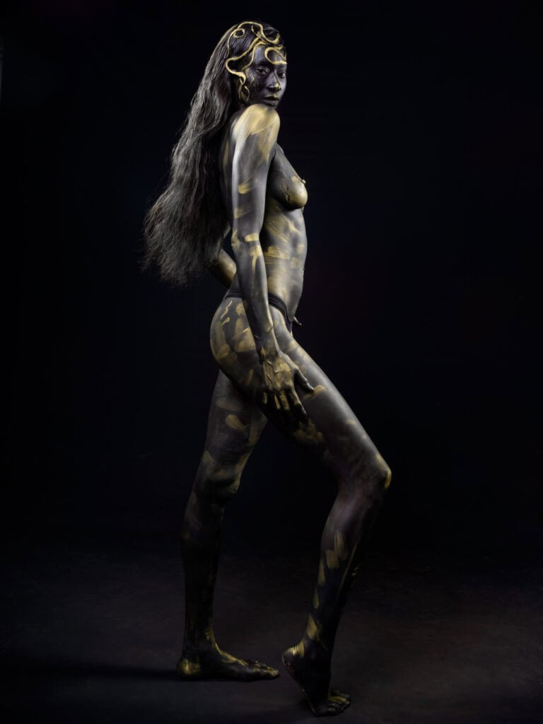 Modèle nu avec body paint doré et noir sur fond noir.