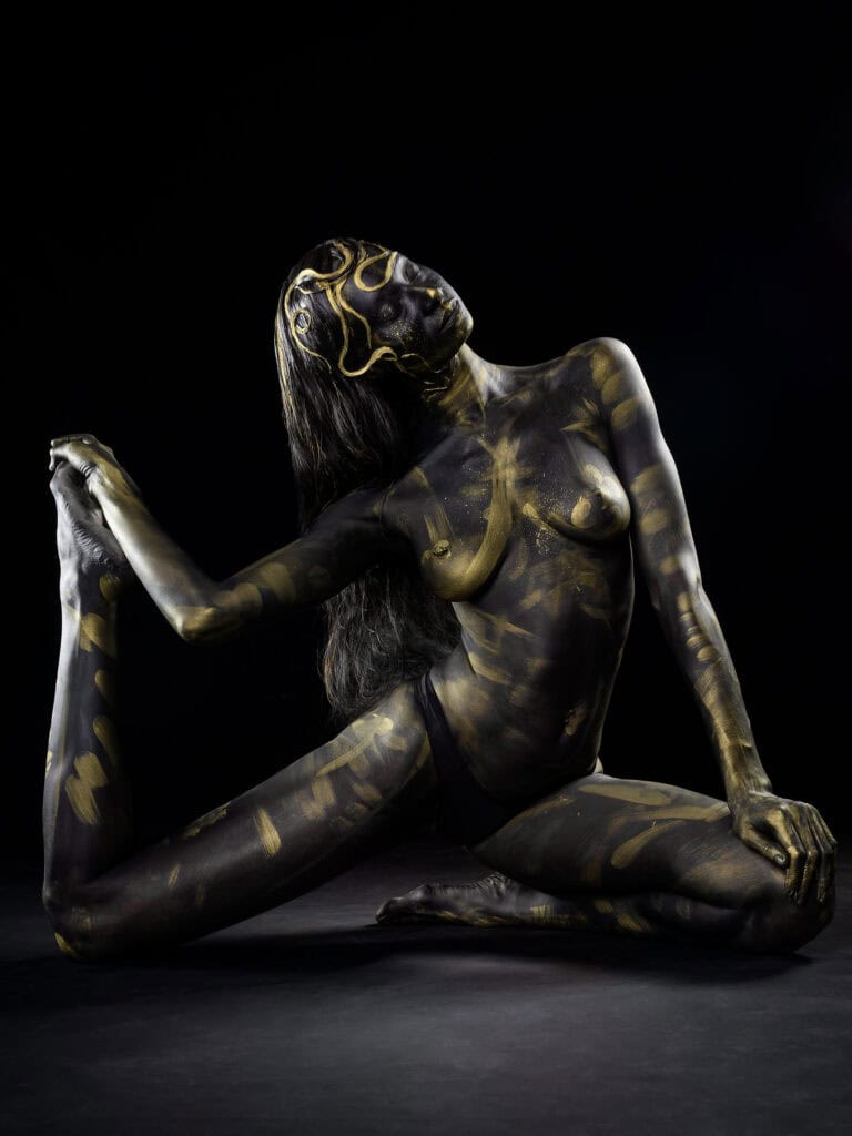 Modèle nu peint en noir avec motifs dorés, pose artistique sur fond noir.