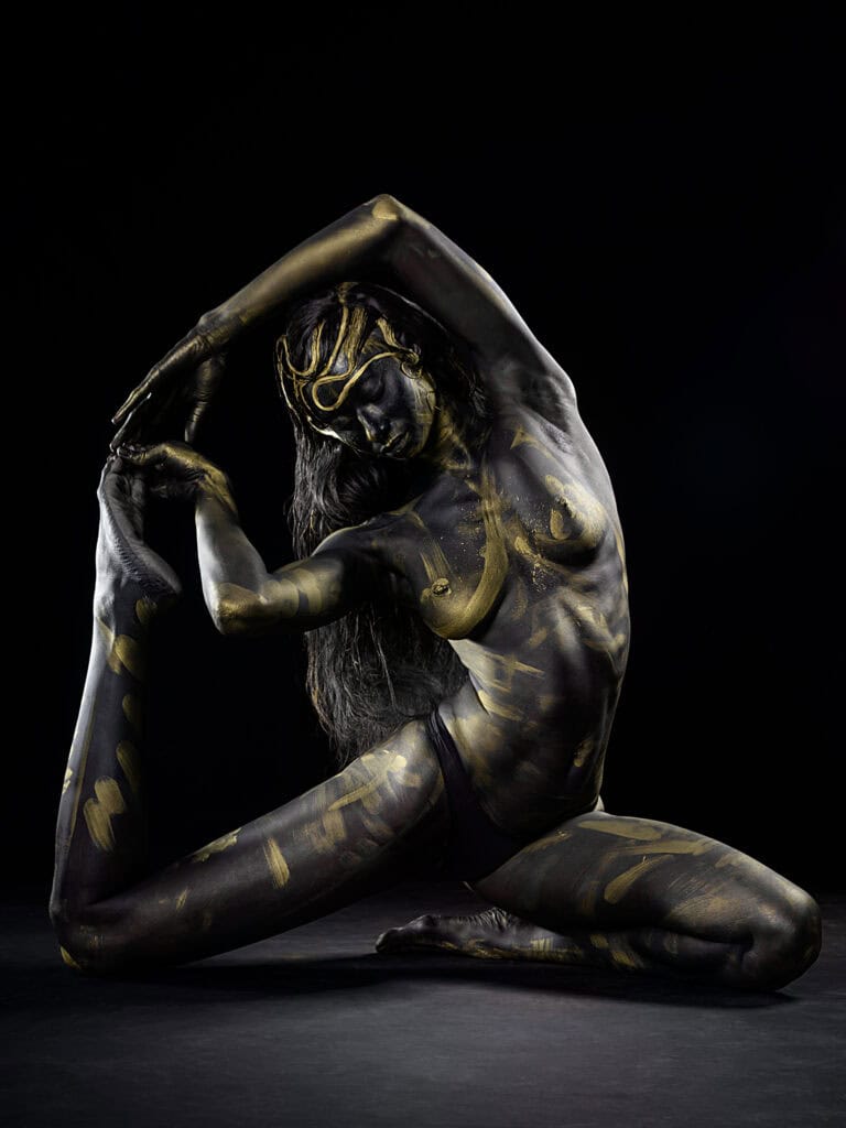 Danseuse en body peinture noire et or, pose artistique sur fond noir.