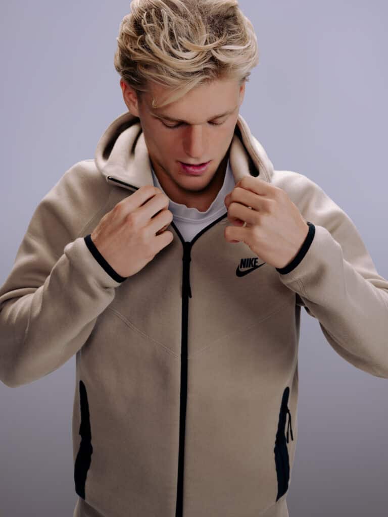 Jeune homme en veste Nike beige, regardant vers le bas, fond gris clair.